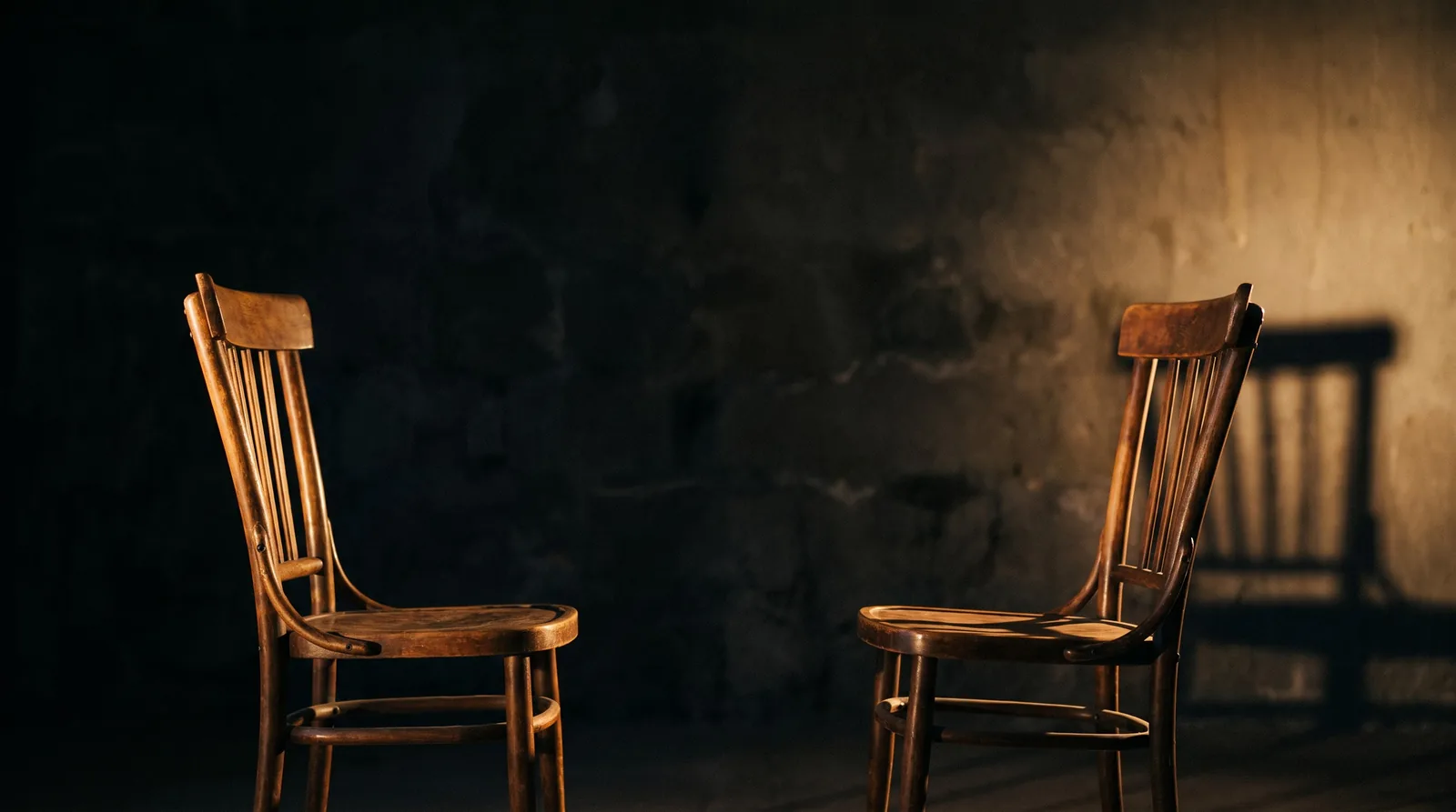 Two empty chairs facing each other in a bare room, single warm light source